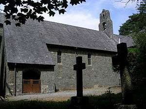 Part of a stone church with a large slate roof seen from a slight angle; on the left is a slightly protruding porch with wooden gates, and on the summit is a bellcote