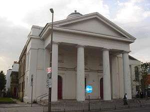 A Classical style, stuccoed building whose façade is dominated by four tapering columns supporting a pediment. The side wall is yellow brick. Partly hidden behind the columns are two red round-headed doors. Above the pediment is a partly hidden cupola. A modern extension is partly visible to the right.