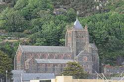 A church seen from the south at a distance with a tree-covered rocky hillside beyond; the church is large with a clerestory, a castellated tower with a small pyramidal spire and clock faces on the two visible sides