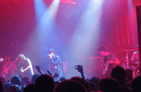 Two guitarists, a drummer, a bassist, and a vocalist are performing a song live on a stage lit by red and blue lights. The crowd, some of whom have their hands in the air, are visible in the foreground.