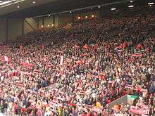 A stand which is full of people standing to support their team. There are a number of flags, scarfs, and banners in the crowd.