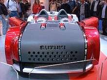 A matte black and bright red two-seat roadster from the front, indoors on a platform with a crowd looking on behind.