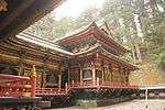 Wooden building connected to a lower building. Both are colored in black and have gilt metal decorations. There is a veranda with red handrail on both buildings.