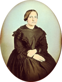 Photographic portrait of a woman seated and wearing a dark dress trimmed in dark lace, with her hair pulled back into a bun and no jewelry except for a simple ring on her left hand