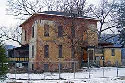 A two-story brick house in extremely poor condition, with boarded-up windows and extensively discolored white paint. It is fenced off.