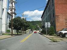 A highway runs toward a bridge between an ornate building with a clock tower and a plain three-story red brick building. A forested hill rises on the far side of the bridge, which is decorated with U.S. flags on poles. A double highway sign along the road indicates that Highway 120 turns to the left (west) before the bridge and that Highway 664 continues across the bridge and heads generally north. Several cars are moving along the highway and over the bridge.