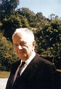 Outdoor photograph of an older man with thinning white hair, dressed in a suit.