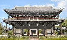 A large two-storied wooden gate with white walls, a veranda on the upper story and a hip-and-gable style roof. Two small wooden open structures with gabled roofs are placed next to it on either side.