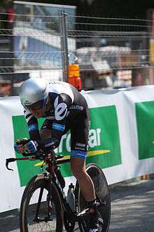 A road racing cyclist in a black and white skinsuit with blue trim and an aerodynamic helmet sits crouched low on his bicycle. There is a guardrail in the background.