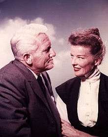 Hepburn is sitting with Spencer Tracy, she age 50 and he age 57, and they are smiling at each other.