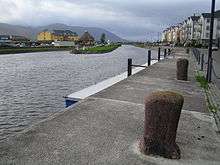 Ship canal and quay, bordered by houses and a paved road. On the horizon in the distance, a high mountain ridge can be seen.