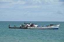 Seven or eight people work to load logs from a smaller, wooden vessel (carved from a tree trunk) onto a slightly larger boat