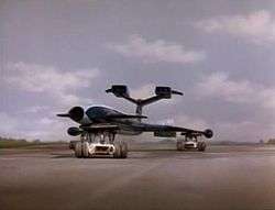 A gleaming, blue, futuristic airliner rests on top of three orange support vehicles (one underneath the fuselage and two underneath the wings) as it speeds out of control along a runway against a grassy backdrop, the sky bright with clouds.