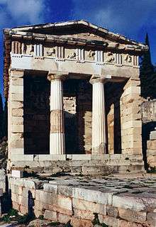 A partially ruined marble building with a porch with 2 columns supporting a pediment and an open doorway beyond