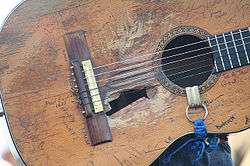 A classical guitar. There are several damages in the soundboard, near the sound hole there is a big hole and the wood is worn out in the surrounding areas of it. The guitar has several signatures on it. there is a blue and white strap in the soundhole.