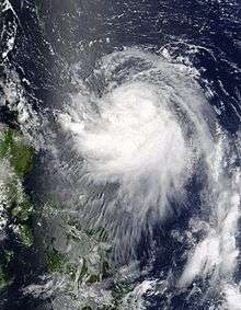 Image of a tropical cyclone to the right of a group of small landmasses. The tropical cyclone is characterized by a sprawling mass of spiralingclouds, though no eye is evident.