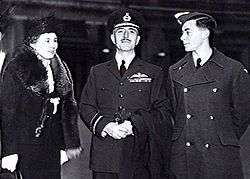 Three-quarter outdoor portrait of moustacioed man in military uniform with peaked cap and pilot's wings on left breast pocket, flanked by woman in hat and fur coat, and young man in military great coat and forage cap