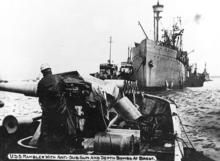 A photograph of two ships. In the foreground is the open bow of USS Rambler with an anti-submarine gun mounted to the left of the frame. USS Bridgeport is in the background of the photo with the bow pointing towards Rambler. There are four smaller ships beside Bridgeport, two on each side.