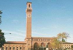 A brick building with a tall clock tower. Its two-story middle section has three tall round-arched windows and a peaked roof, with two one-story wings. In front is a square with a flagpole, statue and some small shrubbery and trees