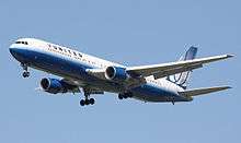 Side quarter view of twin-engine jetliner in flight, with extended gear; white with blue stripes paint scheme.