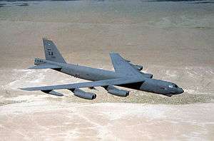 Aerial top/side view of gray B-52H flying over barren desert land.