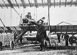 Pilot seated in open-framed  biplane, shaking hands with a man in military uniform who wears a peaked cap