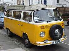 A yellow and white Vokswagen Type 2 vehicle with a dirty roof is parked on a street.