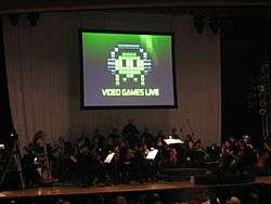 A photograph of an orchestra on a dimly lit stage. Above the group is a projection screen with a black, white, and green image of pixel art. The pixel art is of an oval object wearing headphones with eyes and four tentacles. Below the pixel art is the phrase "Video Games Live".