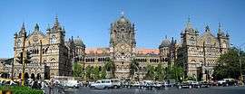 A brown building with clock towers, domes and pyramidal tops. Also a busiest railway station in India. A wide street in front of it