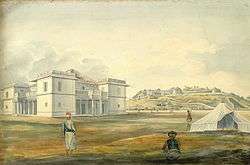 A white mansion and, in the distance, a fortified palace atop a hill. Three men stand near a tent in the foreground. The mansion has a large rust-colored shingled roof, pillared porches and open-air balconies on its four sides, and large box-like corner sections that extend out from the rest of the architecture.