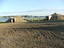 Two dirt-covered earth lodges, with a wooden platform between them and a lake in the background