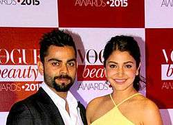  Anushka Sharma, dressed in an yellow dress, and Virat Kohli, dressed in a suit, stare at the camera.