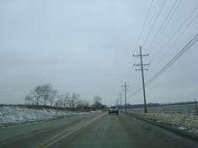 Westbound IL 120 just west of city of McHenry and narrowing to two lanes. Riding on concrete, there are open areas on both sides of the road with scattered trees on the left and utility poles on the right.