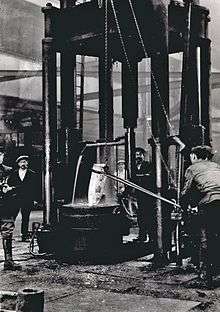 Workers stand around a large forge, where a shell burns red-hot.