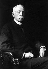 Old white-haired man with white bushy moustache in black suit with high collar sits stern face in a leather chair, legs crossed, staring at a distant point.