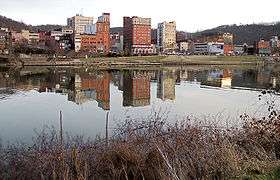 Modest skyscrapers, their images reflected in the water, line the bank of a wide placid river.