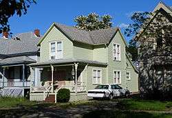 Photograph of a modest two-story wooden house with porch, closely flanked by other houses.