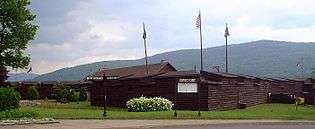 The reconstructed fort is a wooden log construction, painted brown, roughly a single story tall. There are four flagpoles, from which fly a variety of flags, including the American flag and a British Union Jack.  Mountains are visible in the background.