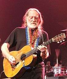 A man with long white hair and white beard playing a guitar. He wears a black T-shirt, which is crossed by the red, white and blue strap of the guitar. He also wears black pants.