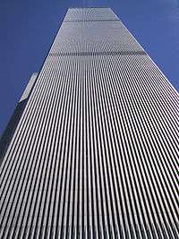 Aerial view of two 110-story twin towers; the building have gray, steel exteriors, and the structure on the left is topped by a large antenna. Several skyscrapers are visible surrounding the two towers.