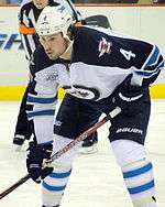 A Caucasian hockey player. He is wearing a white jersey with blue shoulders with a jet over a maple leaf inside of a circle for the logo. He is bent over at the waist leaning on his stick.