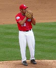 A man wearing a red baseball jersey and cap and white baseball pants standing on a grass mound