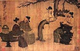 Four men dressed in robes and black square cut hats gather around a tree talking to one another. Three are sitting on rocks while the fourth is leaning over a horizontally bent branch of the tree.