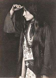 Photo of Zitkala Sa in profile, wearing Native American dress, with long dark hair hanging below waist, holding hand at forehead and looking into the distance