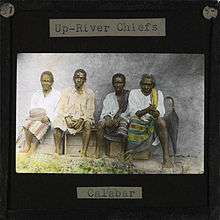 Group picture of four village chiefs visiting Okoyong. The four chiefs, all men, are sitting on a wooden box in front of a clay wall. To the right of the men a black umbrella rests against the wall. Mary Slessor, the Scottish Missionary, lived in Okoyong (located in later day Nigeria) for many years.