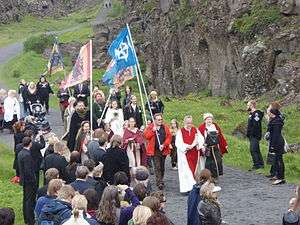 A crowd of people walking along an outdoor path. They are led by individuals in robes and a number carry flag banners.