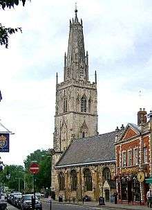 A stone church in a town seen from the southeast.  St the far end is a large tower surmounted by a truncated spire with pinnacles and, at the top, a ball finial