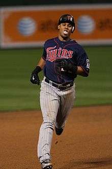 A man in a blue baseball jersey, gray pinstriped pants, a batting helmet, and black gloves runs the bases.