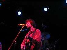 A man with an orange shirt singing to a microphone and playing the guitar.