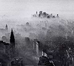 A black-and-white, panoramic view of New York City as seen from a great height. A vast number of buildings and skyscrapers can be seen. A hazy, smoky gas overlays the entire city like a blanket, with a fairly clear skyline only in the far distance at the horizon. Near the closest buildings, the smog appears thin and wispy. The smog appears thicker and thicker around buildings that are farther away from the photographer's position, until shorter buildings near the horizon are almost entirely shrouded and impossible to see under a thick layer of smog. Near the horizon, the clustered tops of tall skyscrapers emerge from within the smog.